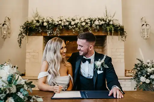 Bride and groom signing the register with floral arrangements adorning the fireplace behind them at a Cotswolds wedding.