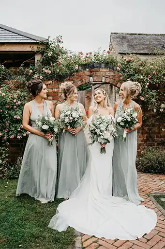 Bride and bridesmaids holding white and green floral bouquets in front of a picturesque brick arch at Blackwell Grange.