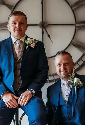 Groom and best man posing in navy suits with blush boutonnières against a rustic clock backdrop.