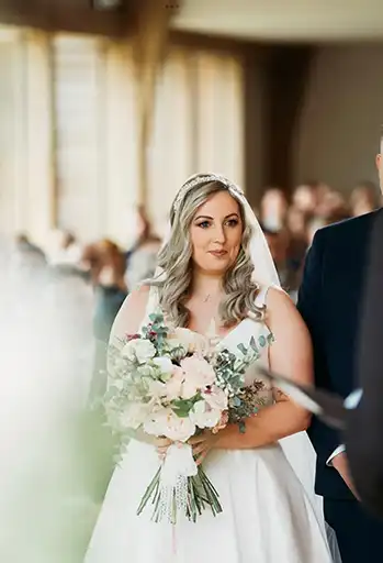 Bride holding a classic bouquet with soft pink and ivory florals, walking down the aisle at The Mill Barns.